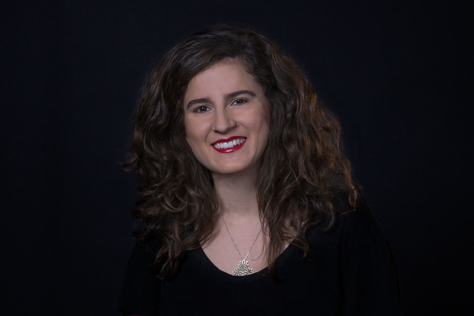 A woman with shoulder-length curly brown hair smiles at the camera against a dark background. She wears red lipstick and a silver necklace with a pendant. Her top is black.