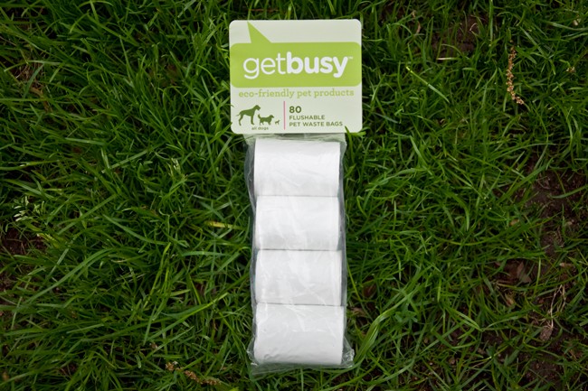 A package of GetBusy eco-friendly pet products lies on a patch of green grass. The package contains 80 flushable pet waste bags and is labeled with the GetBusy logo and text indicating it is pet-friendly and eco-friendly. The package is sealed in clear plastic and rests on the grass.