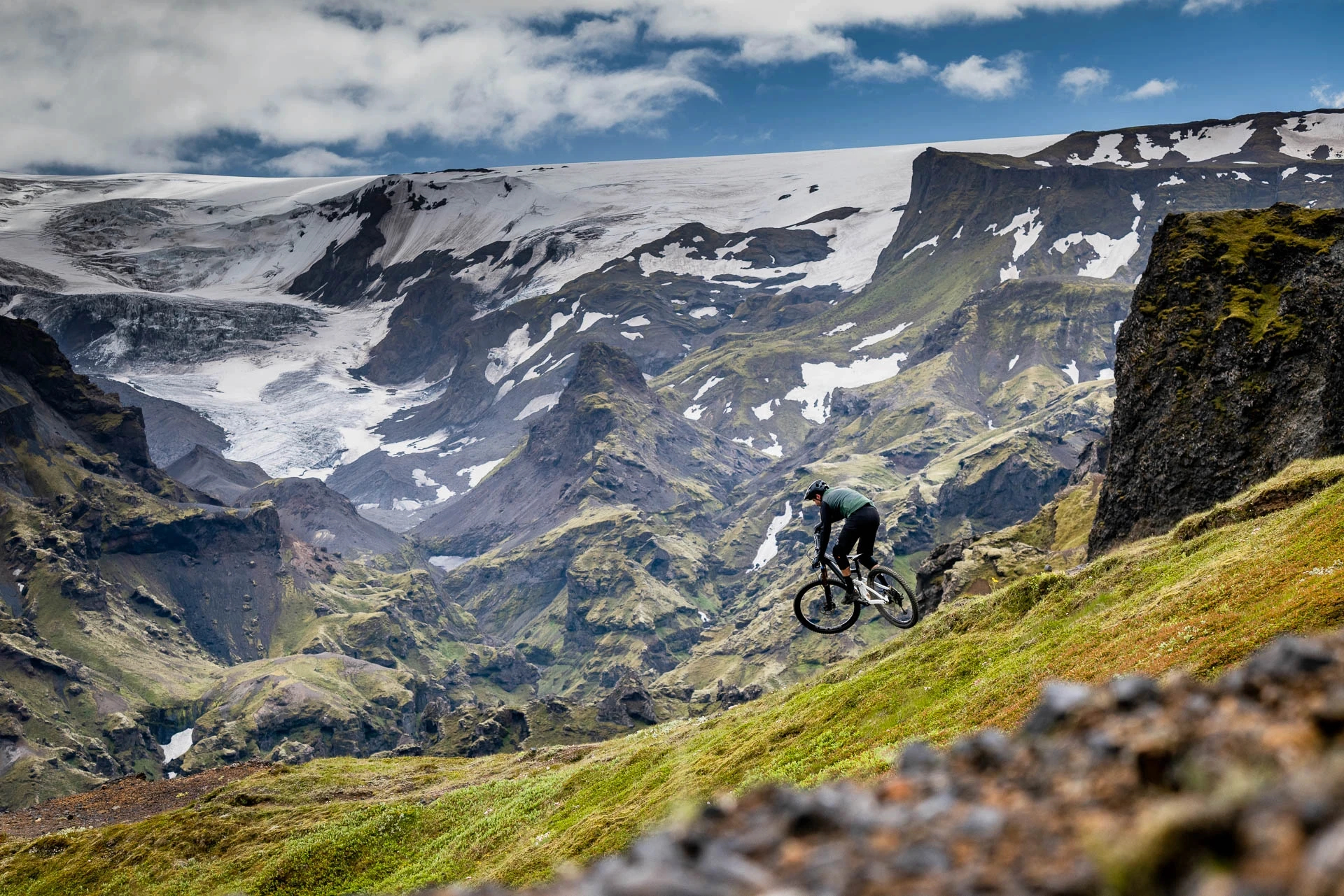 Mountain biker descending a grassy slope with glacier-covered peaks behind.