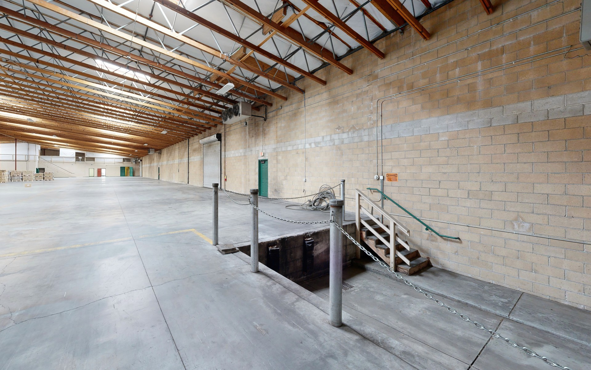 Interior view of a large clear-span warehouse space with polished concrete floors, concrete masonry unit walls, exposed wood truss ceiling with corrugated metal decking and skylights, and a recessed loading pit with chain barriers and metal stairs in the foreground.