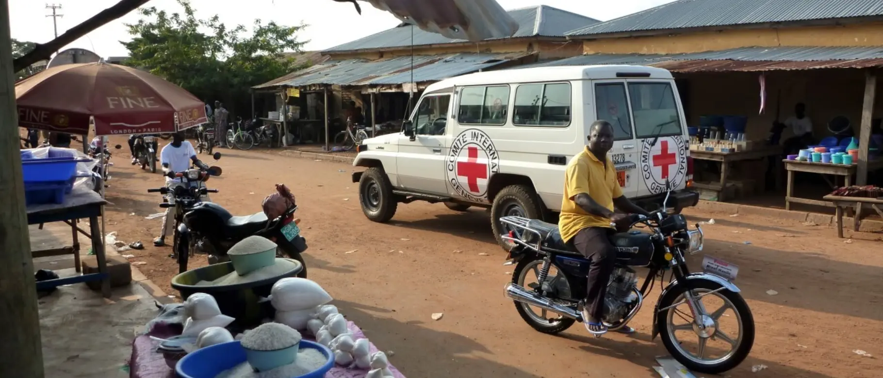 Street scene in Togo with an International Red Cross vehicle, motorcyclist and market stall – representing global connections, supply chains and corporate responsibility.