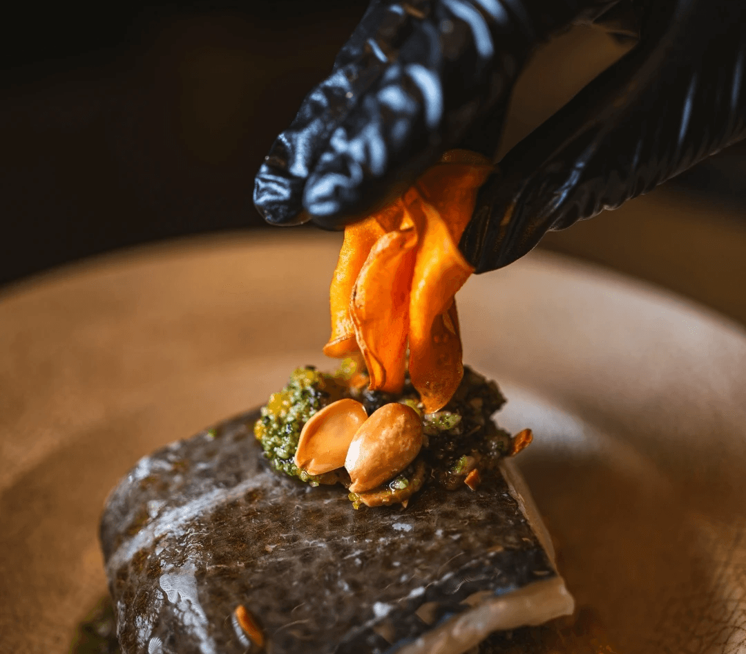 la mano del chef cubierta con un guante negro detallando el emplatado de un plato de pescado