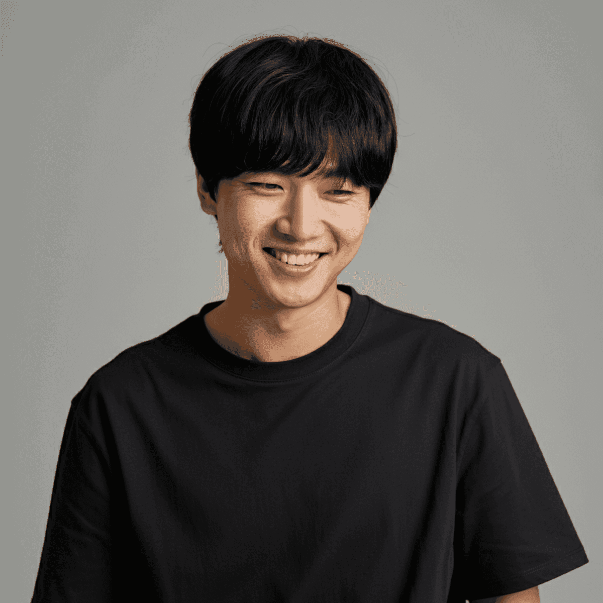 Smiling person with short dark hair in a black shirt against a plain grey background. The expression conveys warmth and friendliness.
