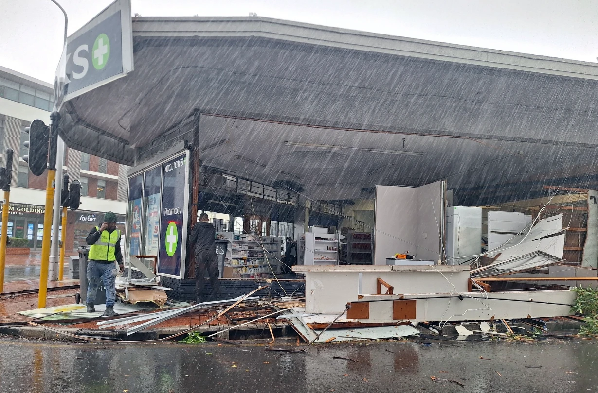 A damaged Clicks store with a collapsed wall as heavy rain falls outside onto the street.