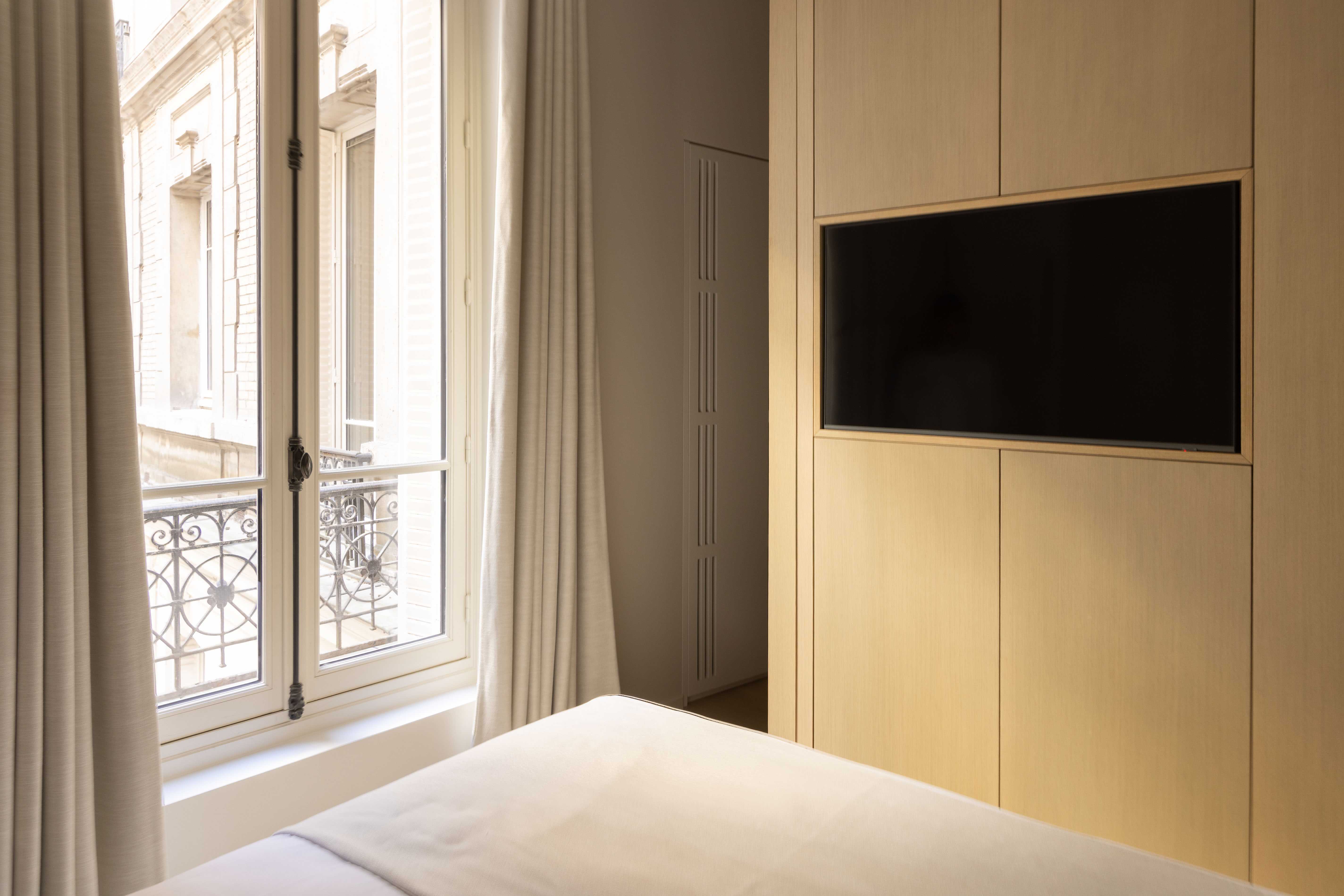 Chambre avec TV encastrée dans meuble bois, fenêtres avec balcon et vue parisienne