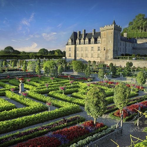 Ornate gardens with manicured hedges and colorful flowers in front of a large historic castle under a clear blue sky.