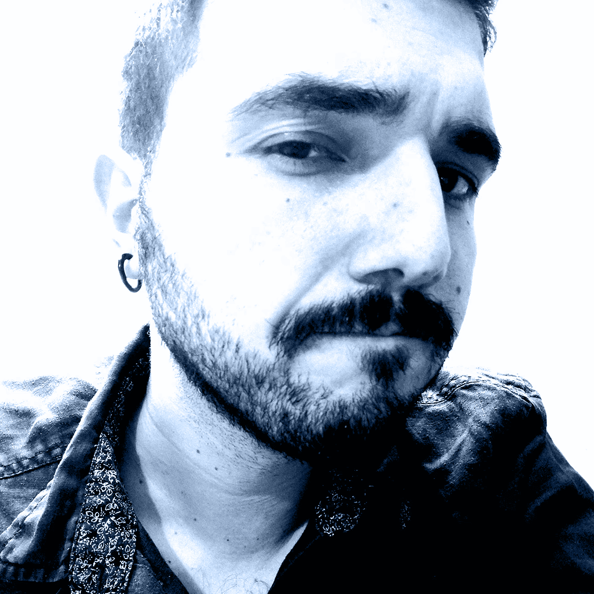 A man with glasses and a beard is looking directly at the camera, seated in a cozy, dimly lit café.