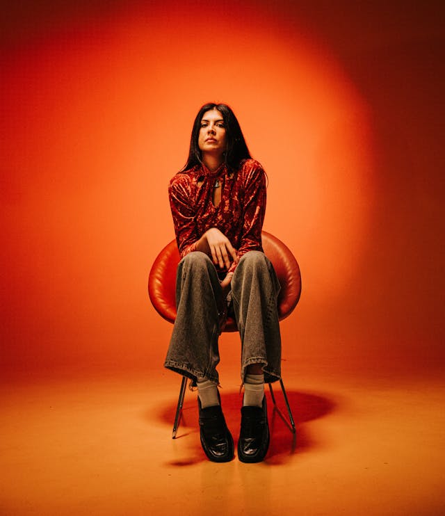 A woman in a patterned shirt and jeans sits on a chair against a solid orange background