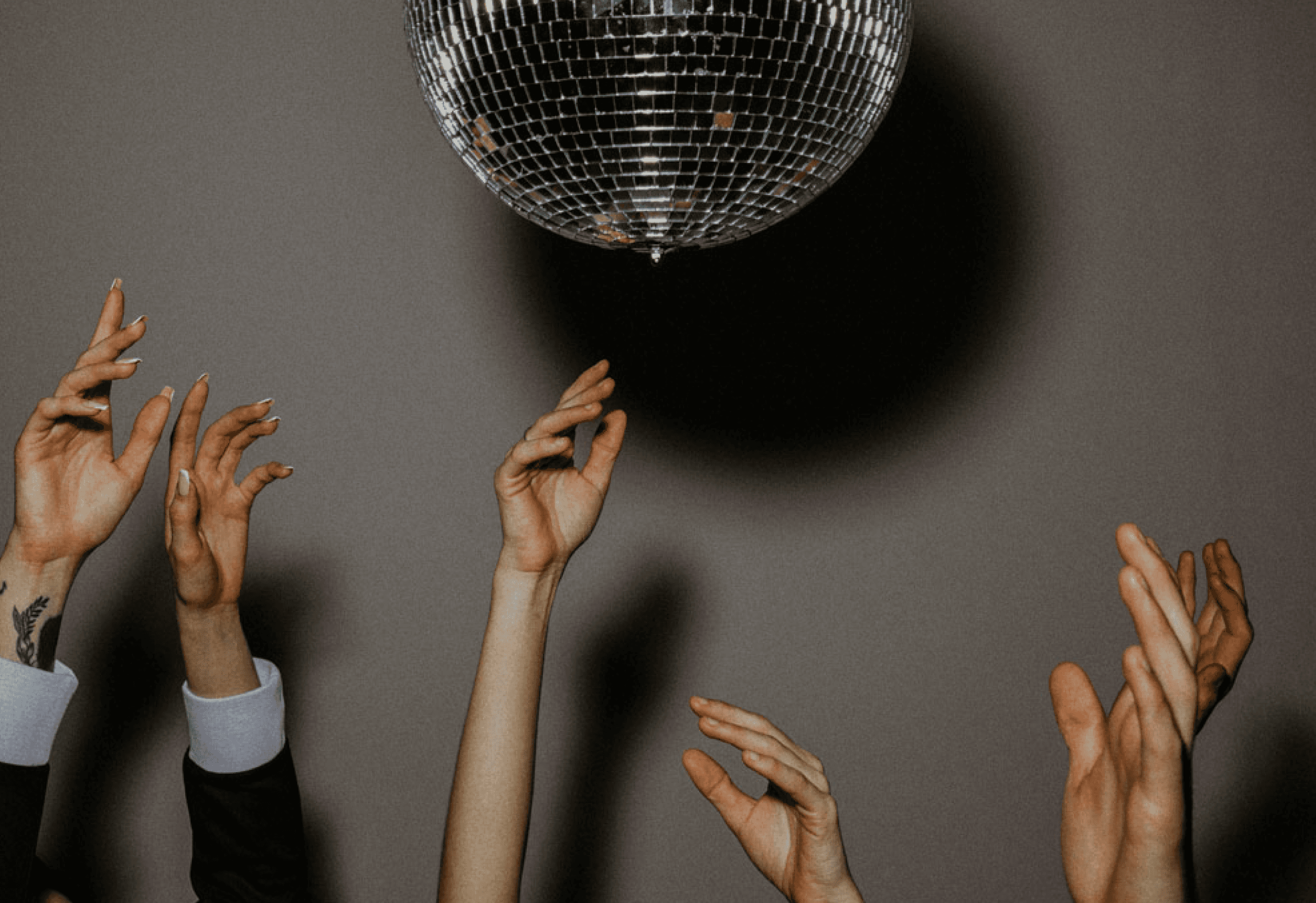 Several hands reaching up towards a large, reflective disco ball hanging in a dark background, creating a dramatic, theatrical composition.