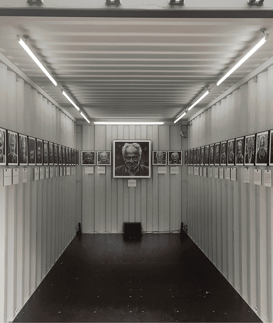 Vue intérieure d’un conteneur d’exposition présentant les portraits en noir et blanc de Frédéric Bourcier, éclairés par des néons au plafond.