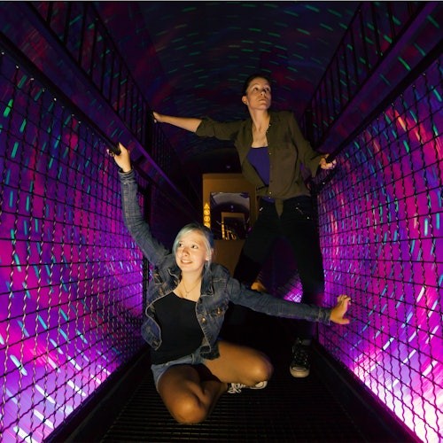 Двама души, един клекнал и един изправен, в тунел с ярко лилаво и синьо осветление и мрежести стени.