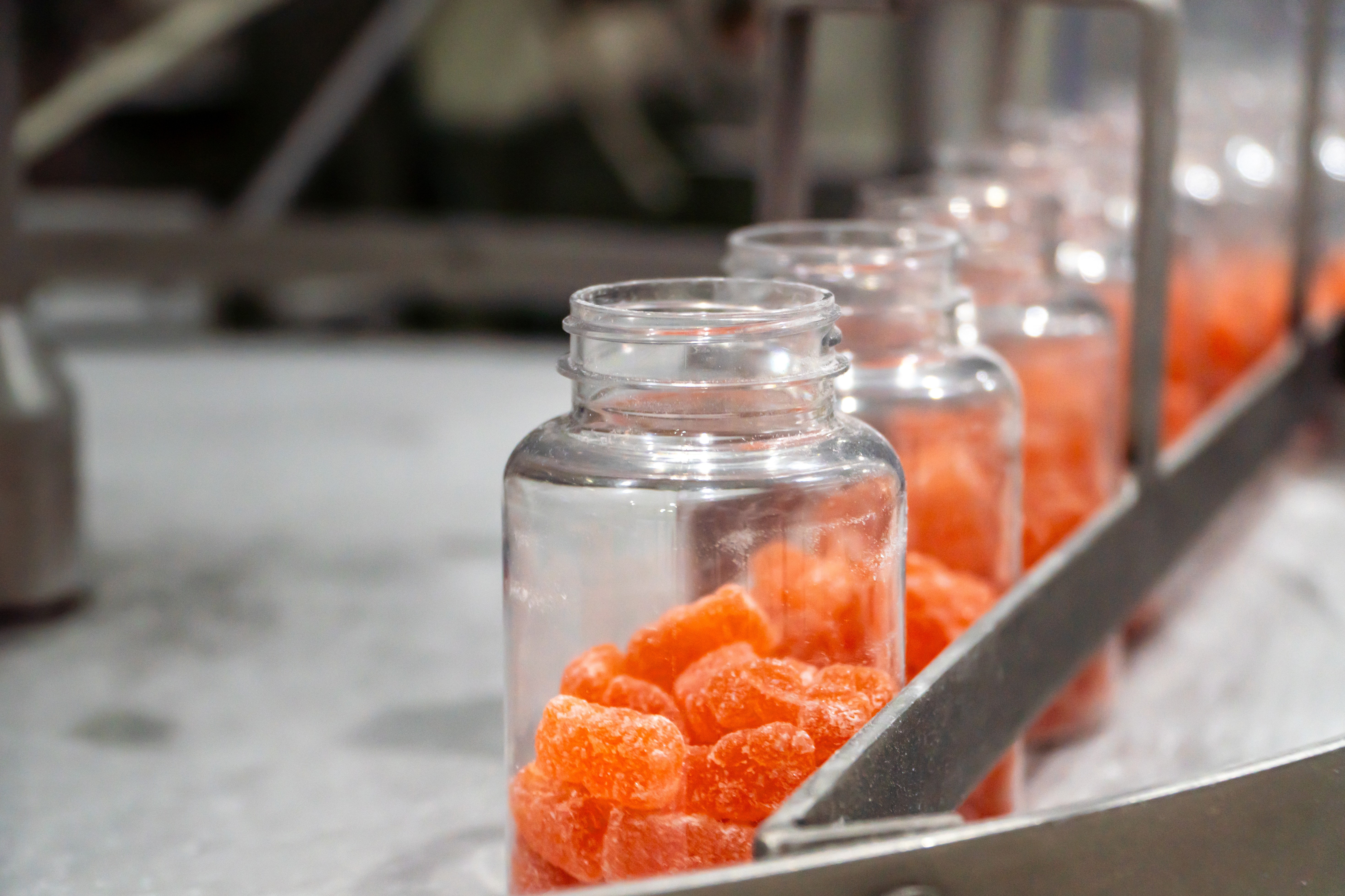 Clear jars lined up in a manufacturing or packaging facility containing red gummies.