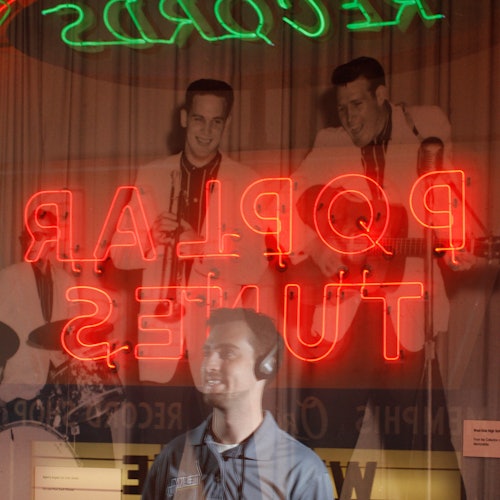 Mladý muž s&nbsp;nasazenými sluchátky stojí před neonovým nápisem a fotografií hudebníků při vystoupení.