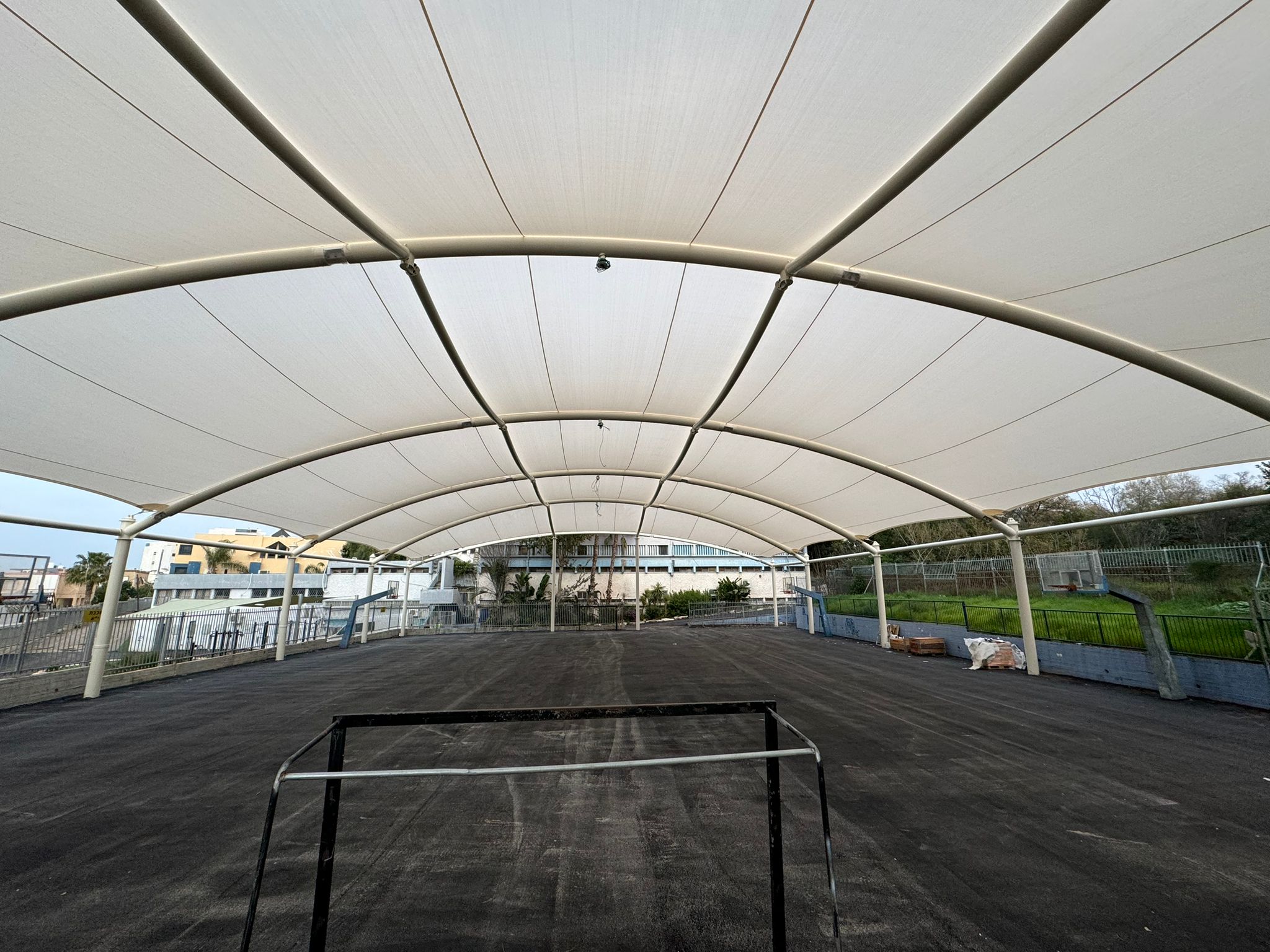 Large covered area with beige tent roof and black floor surface surrounded by railings and buildings in the background.