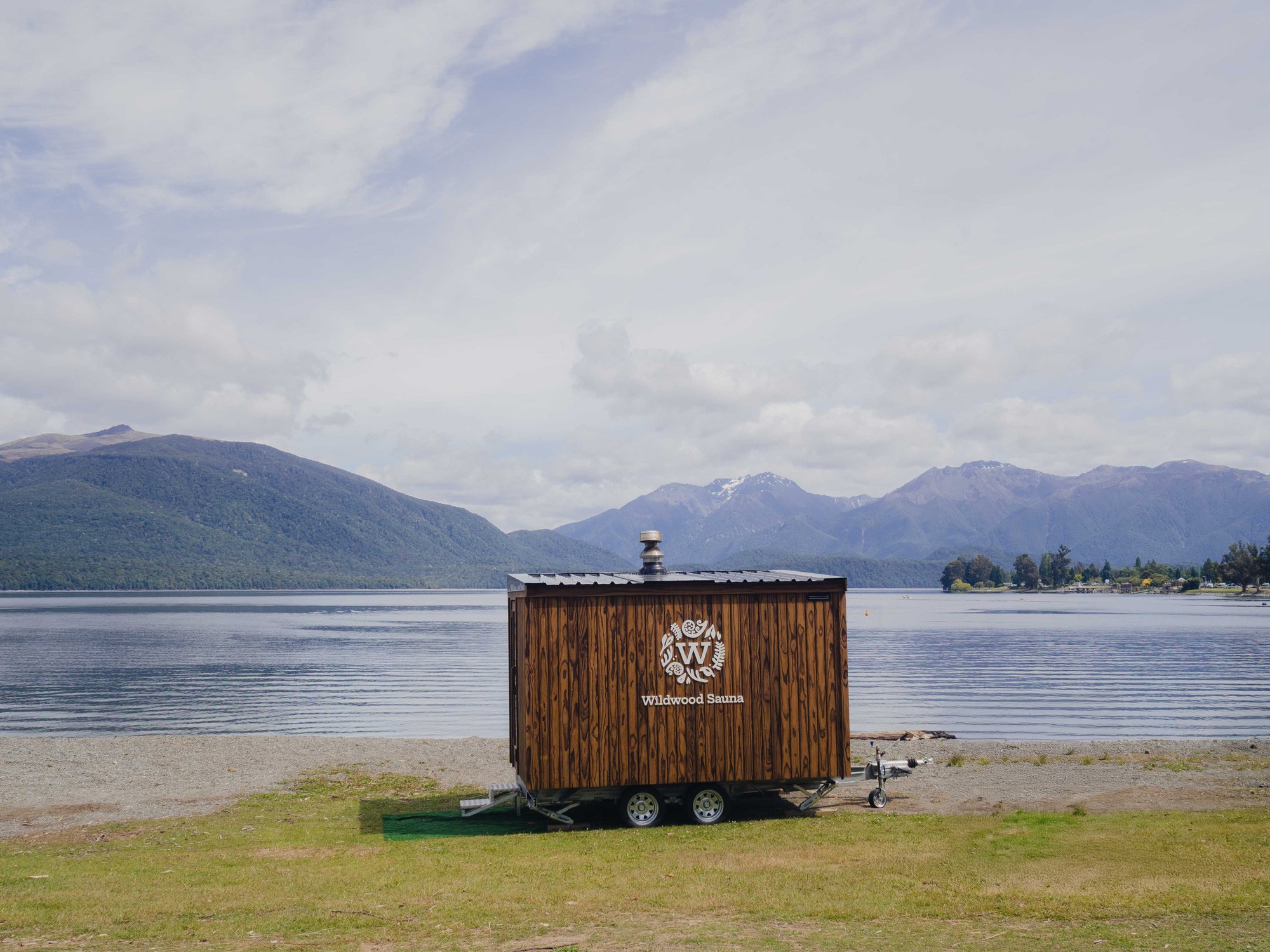 Mobile sauna parked at calm lake overlooking a mountain range.