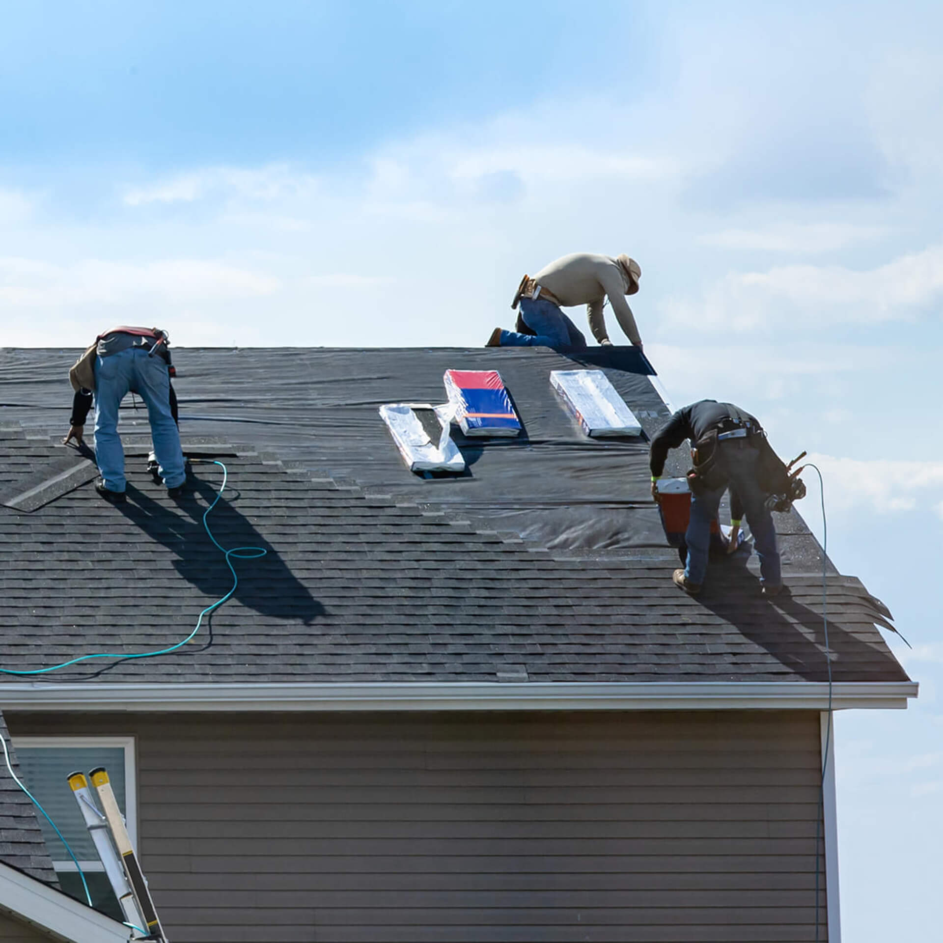 Three workers install asphalt shingles on a residential roof under a clear sky, with roofing materials and tools spread across the surface. The scene shows part of the roof already completed, emphasizing the active progress of a professional roofing job.