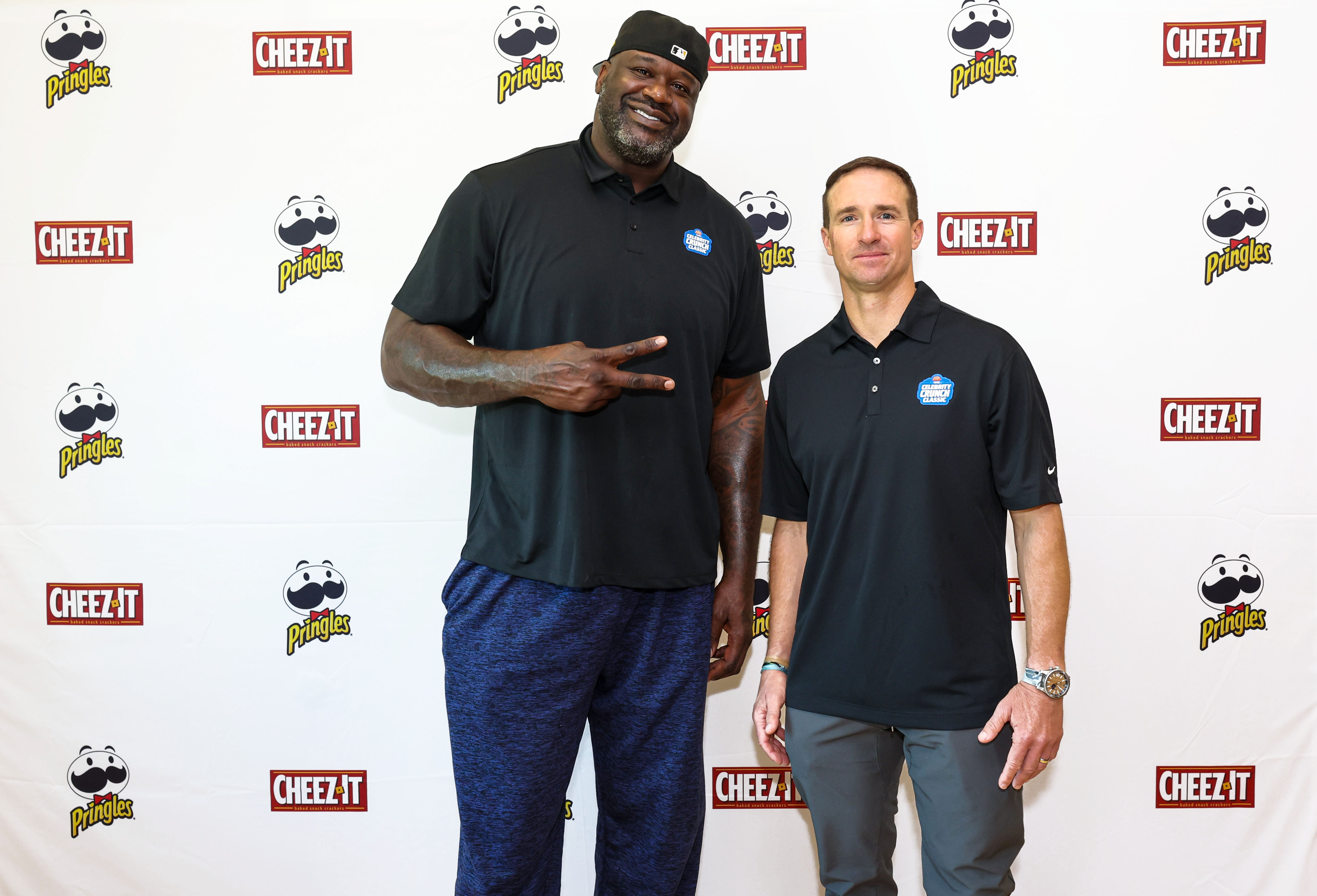 Shaq and Drew Brees in black polo shirts stand side by side, smiling, in front of a backdrop with Cheez-It and Pringles logos. One man is significantly taller and is holding up a peace sign.