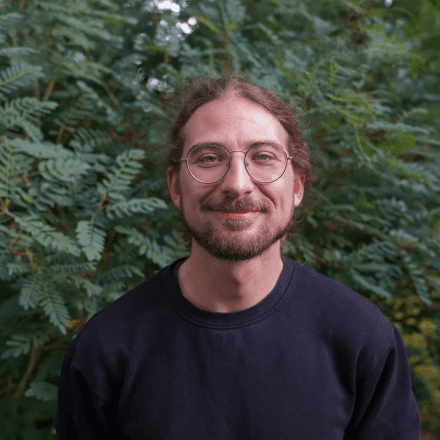 Mann mit Brille und Bart vor grünen Blättern im Freien lächelnd.