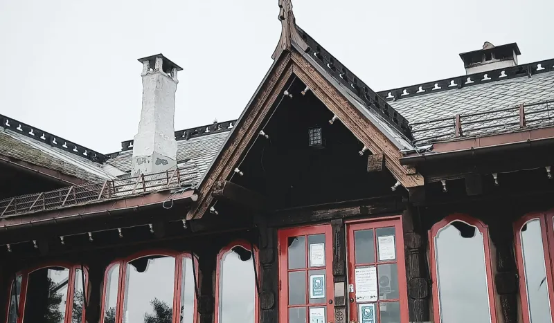 Traditional wooden house exterior with a steep roof, chimney, and red-trimmed windows.