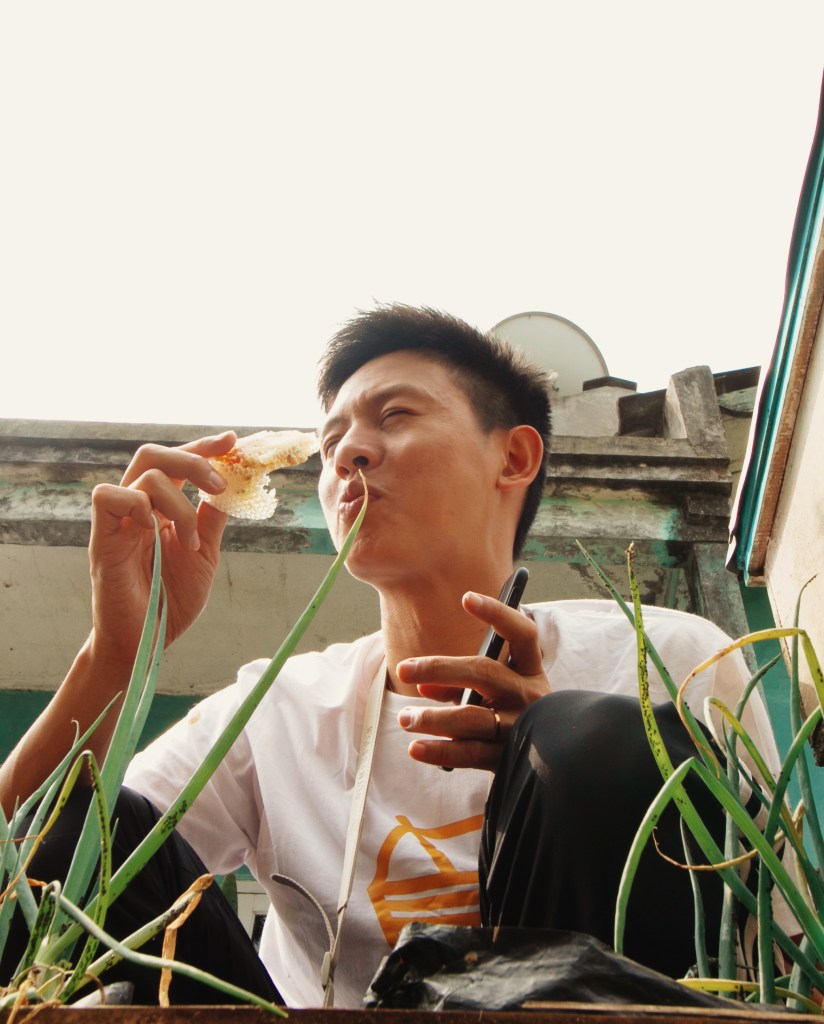 A young man sitting in a garden, enjoying a snack while surrounded by green grass and plants.