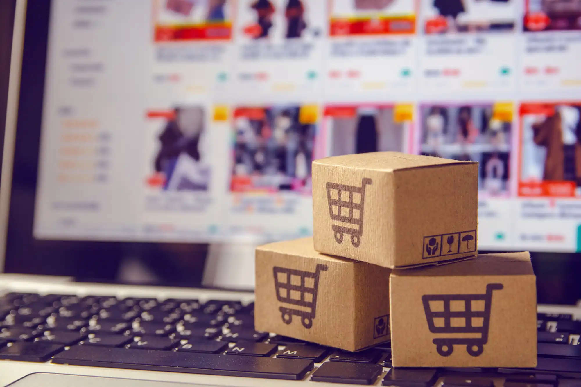 Small cardboard boxes with shopping cart icons sit on a laptop keyboard, representing the growing online retail and shipping industry.