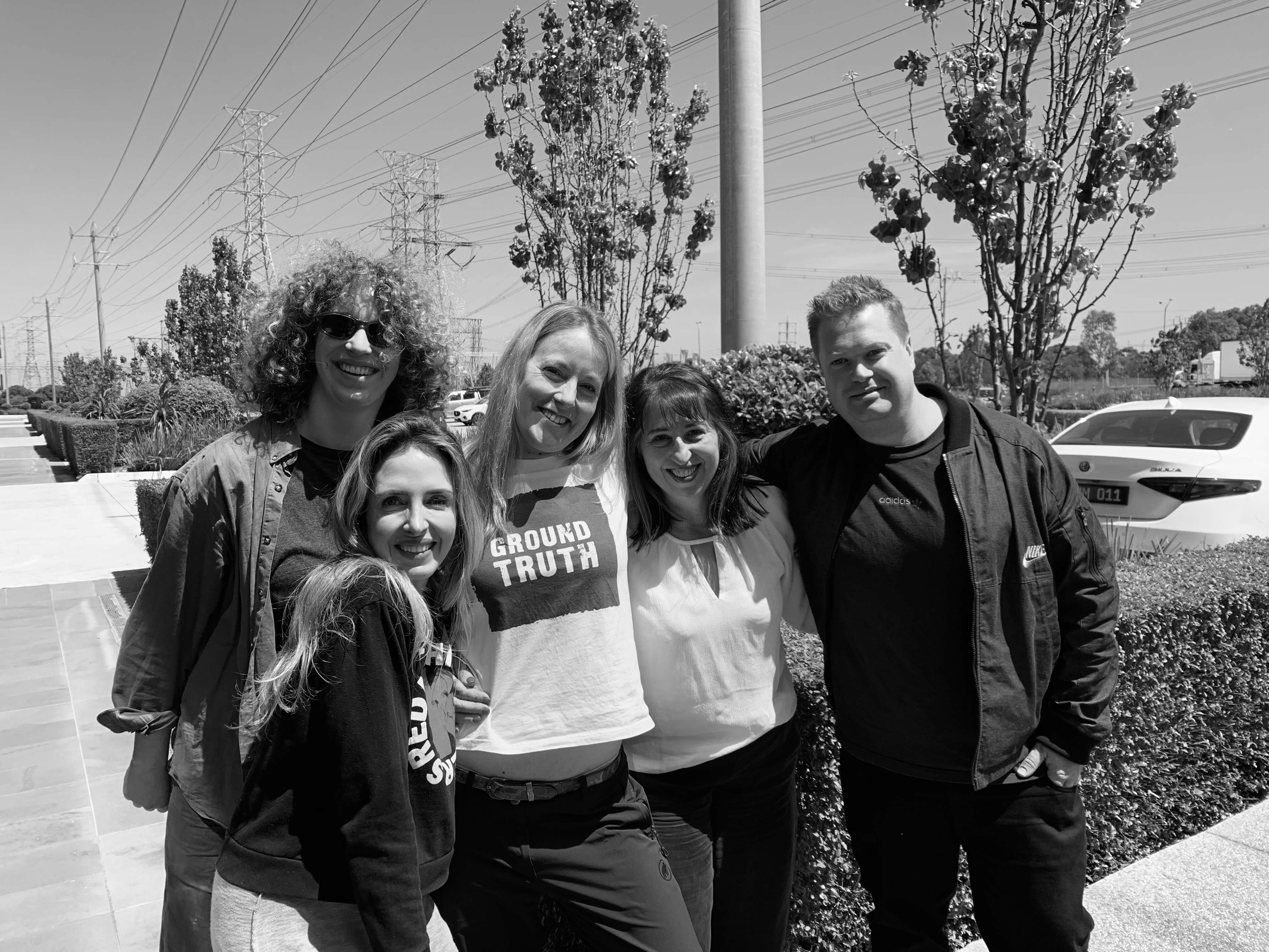 A group of five people smiling together outdoors in black and white, with trees and a building in the background.