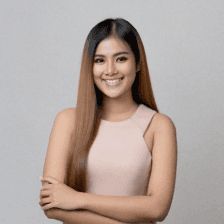 Smiling woman with long brown hair wearing a tan sleeveless top against a neutral gray background.