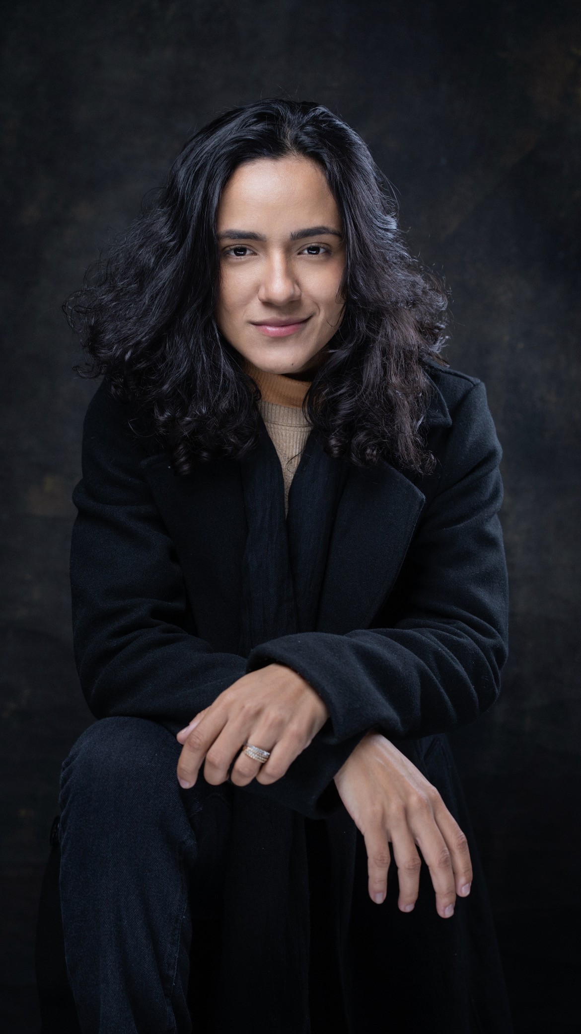 Una persona con una camiseta negra y pantalones de color claro está de pie en la cima rocosa de una montaña al atardecer, sosteniendo una cámara frente a su cara para tomar una foto. Un mar de nubes se extiende detrás de ellos, iluminado por la luz dorada del sol poniente.
