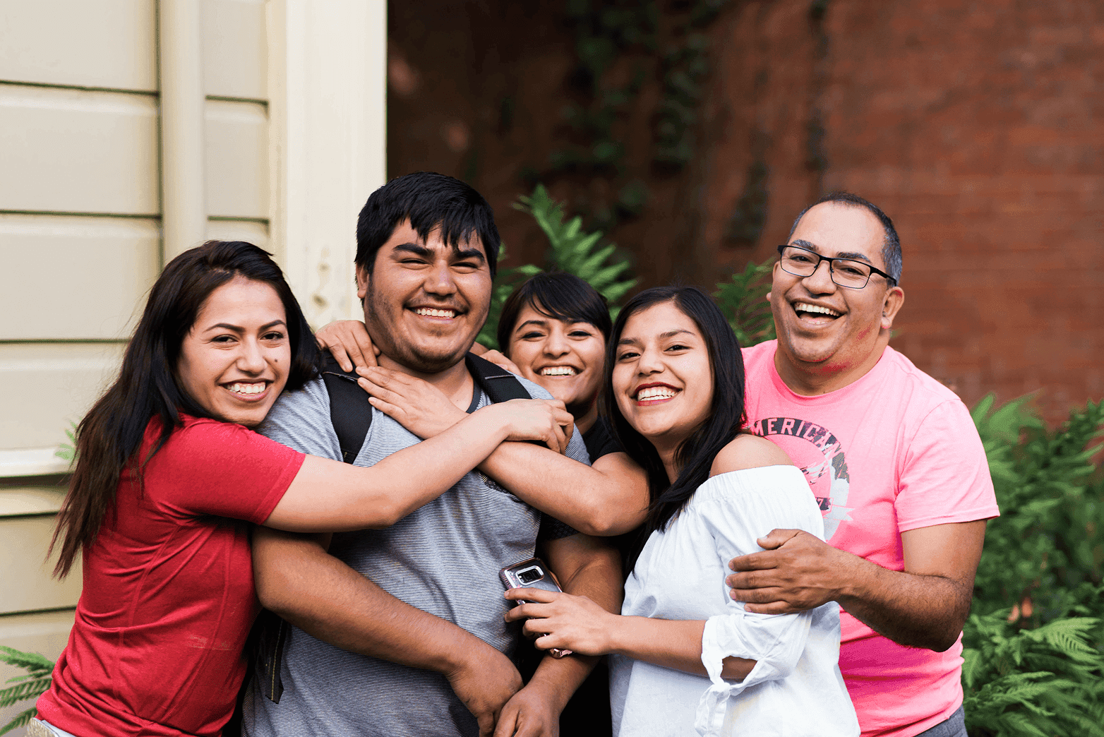 Un grupo de cinco personas se reúne muy cerca al aire libre, sonriendo ampliamente y abrazándose frente a una pared de ladrillo y follaje verde, transmitiendo una sensación de alegría y unión.
