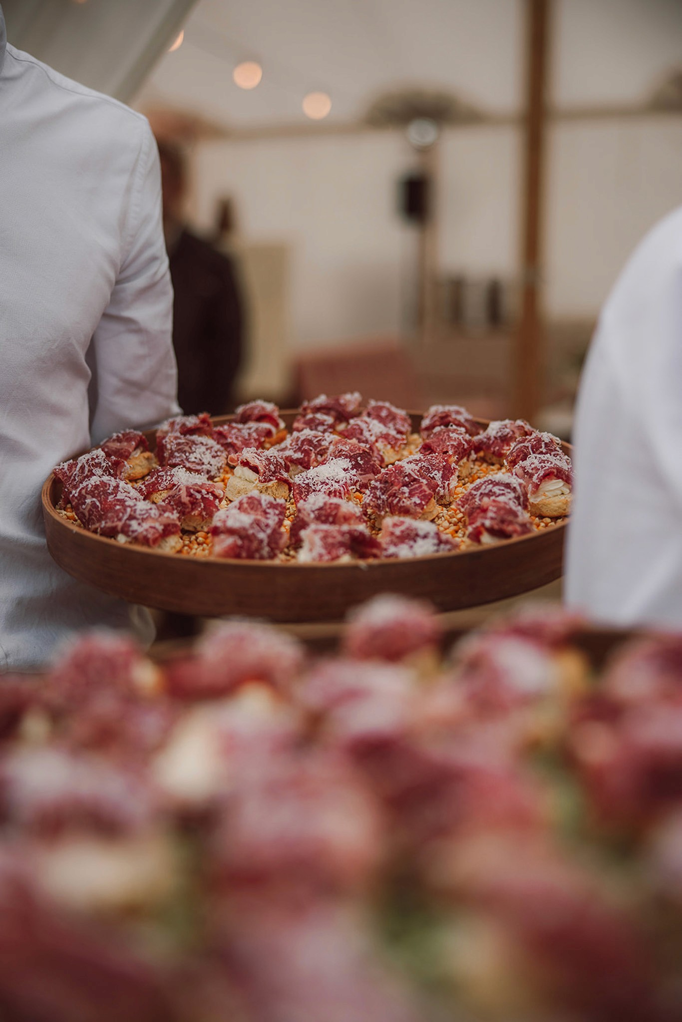 Person with a mustache wearing a black t-shirt carrying a tall stack of pizza boxes inside a dimly lit bar or restaurant.