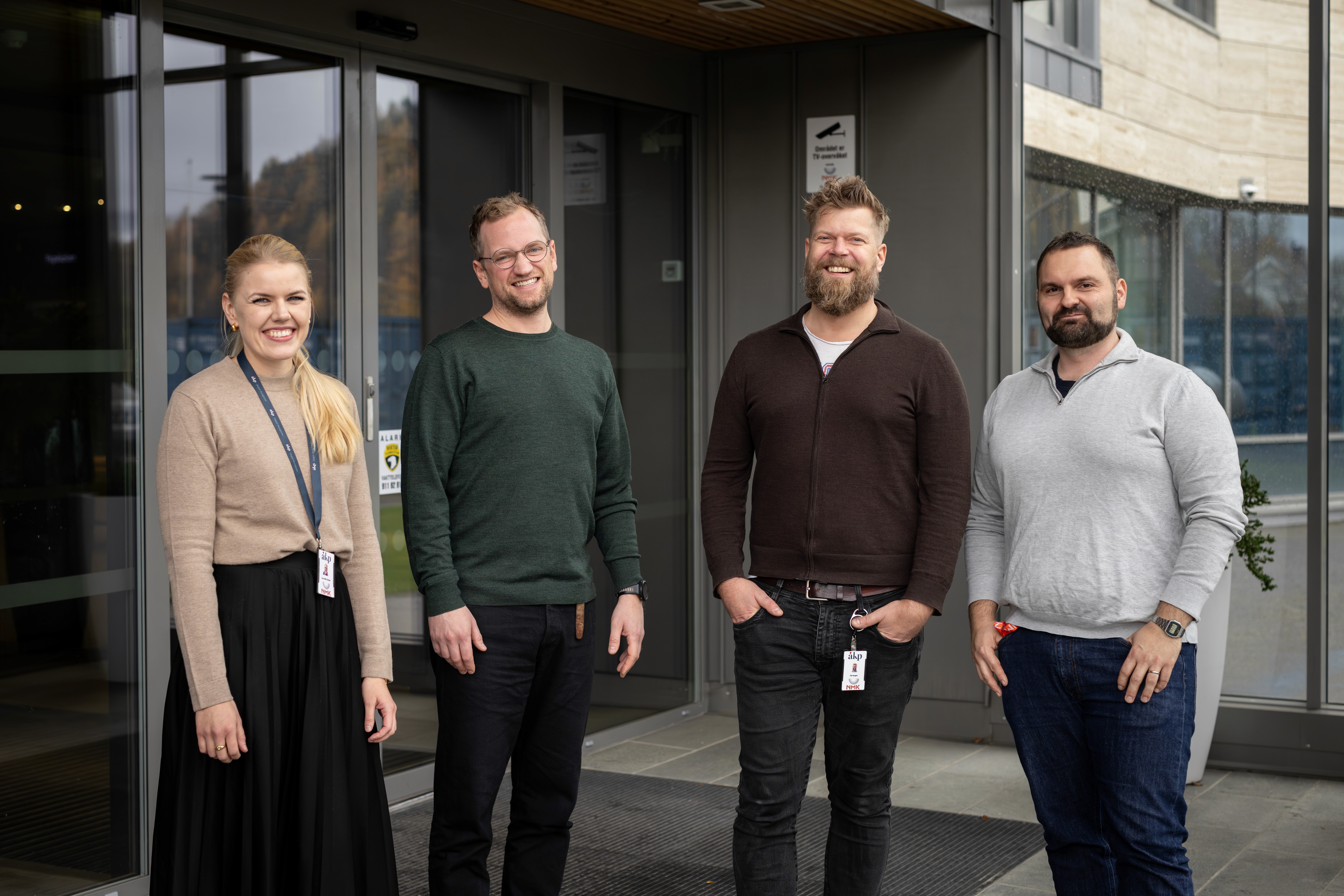 Four individuals stand outside a building, smiling and posing for the camera. They appear engaged and friendly.