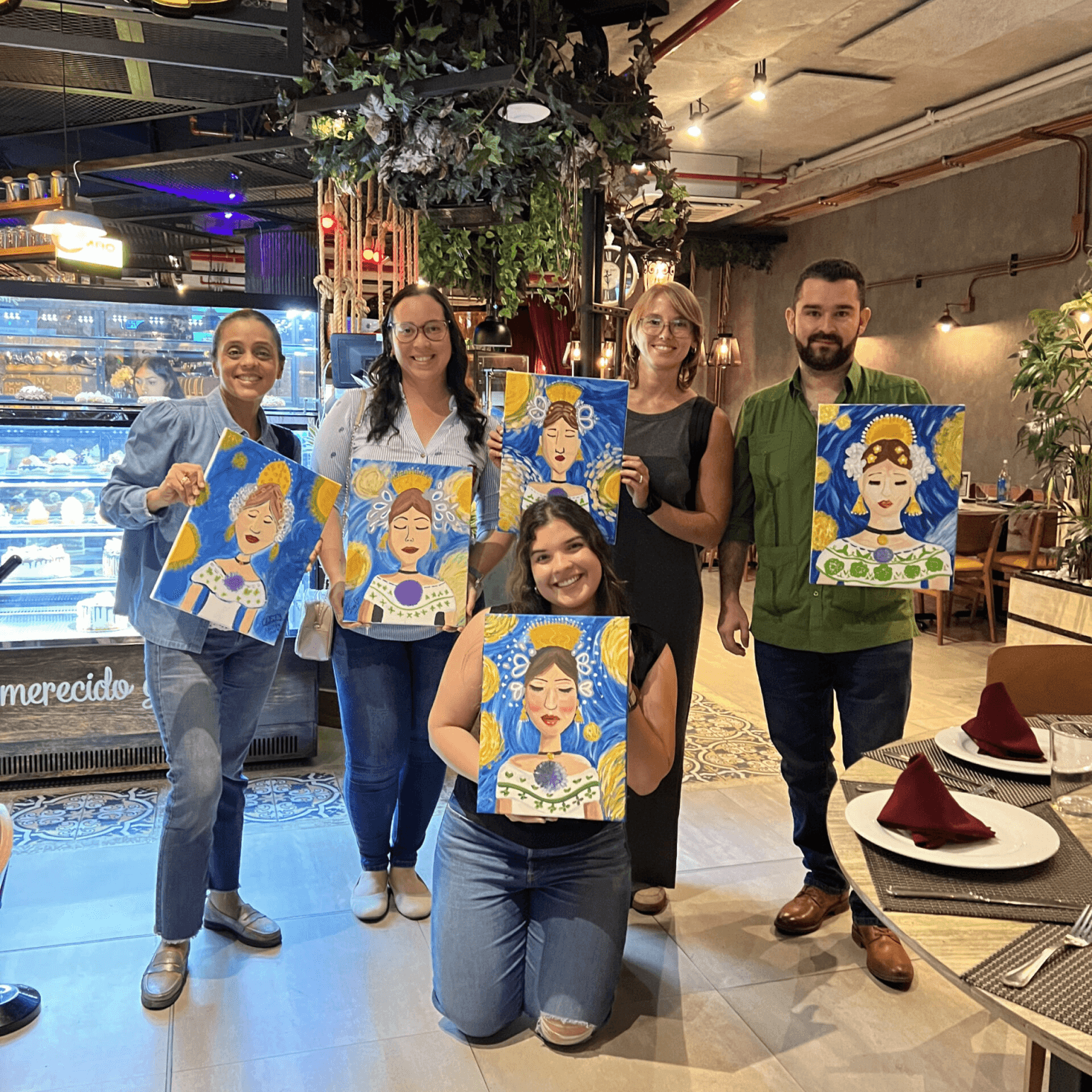 Cinco compañeros posan en un restaurante después de una actividad de pintura, cada uno sosteniendo un colorido retrato en un lienzo azul y amarillo.