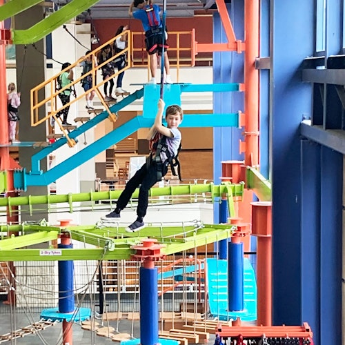 Children in harnesses navigate through an indoor ropes course with colorful platforms and safety structures.
