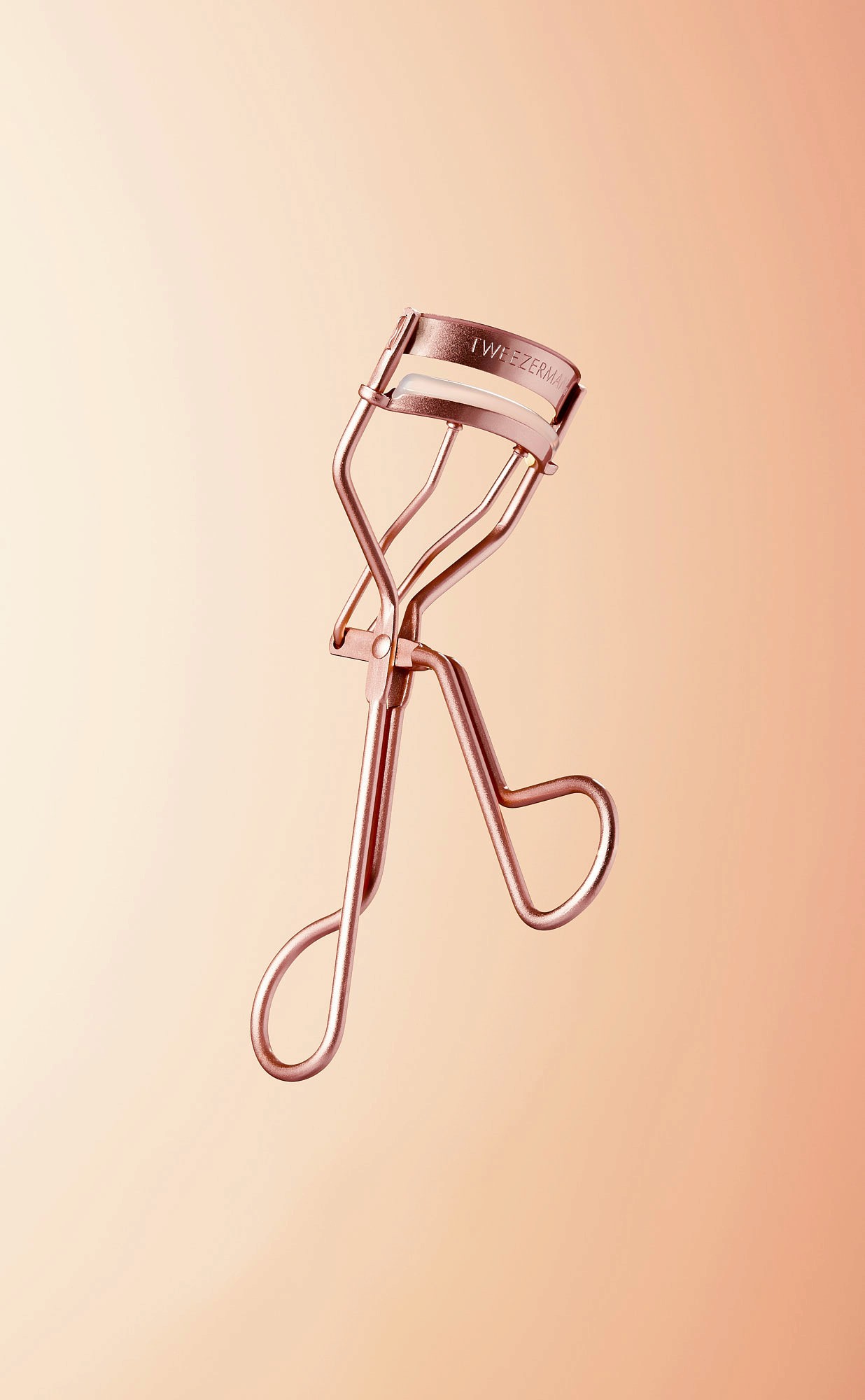 A rose gold eyelash curler against a soft peach background.