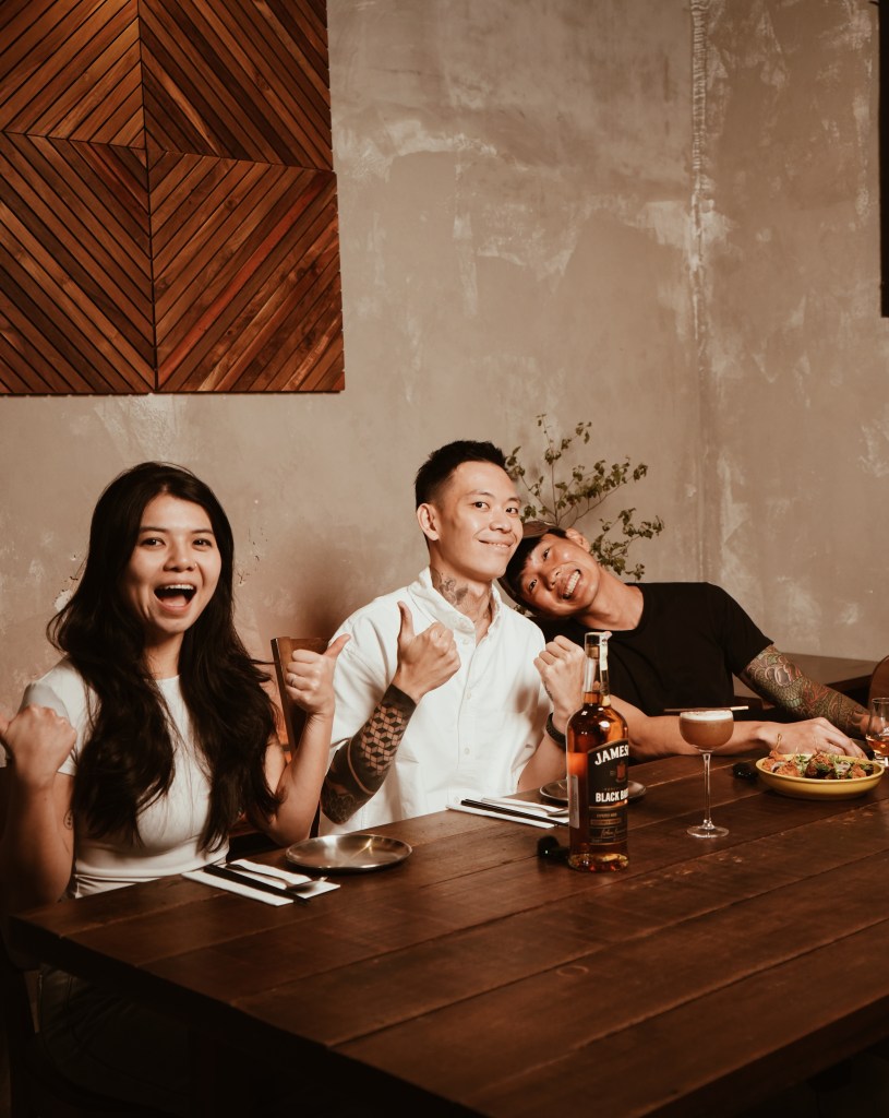 Three people enjoying a festive meal at a wooden table with drinks and food, featuring a bottle of Jameson Black Barrel whiskey prominently displayed.