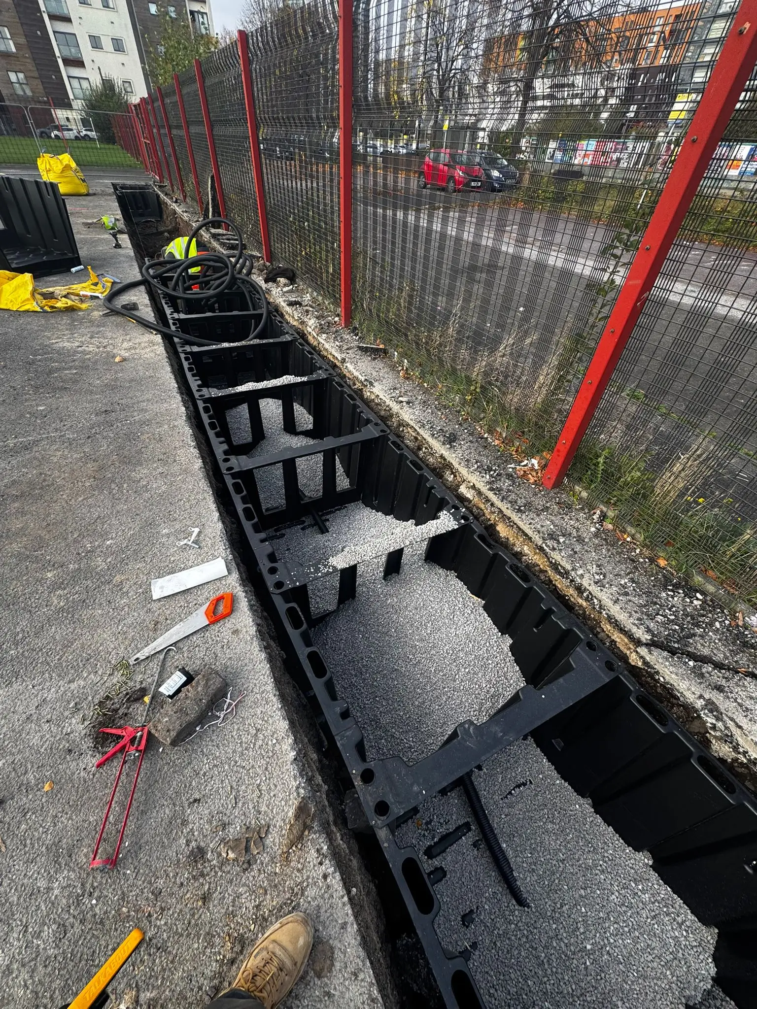 A narrow trench alongside a road, revealing black pipes and construction tools nearby, with red safety fencing.