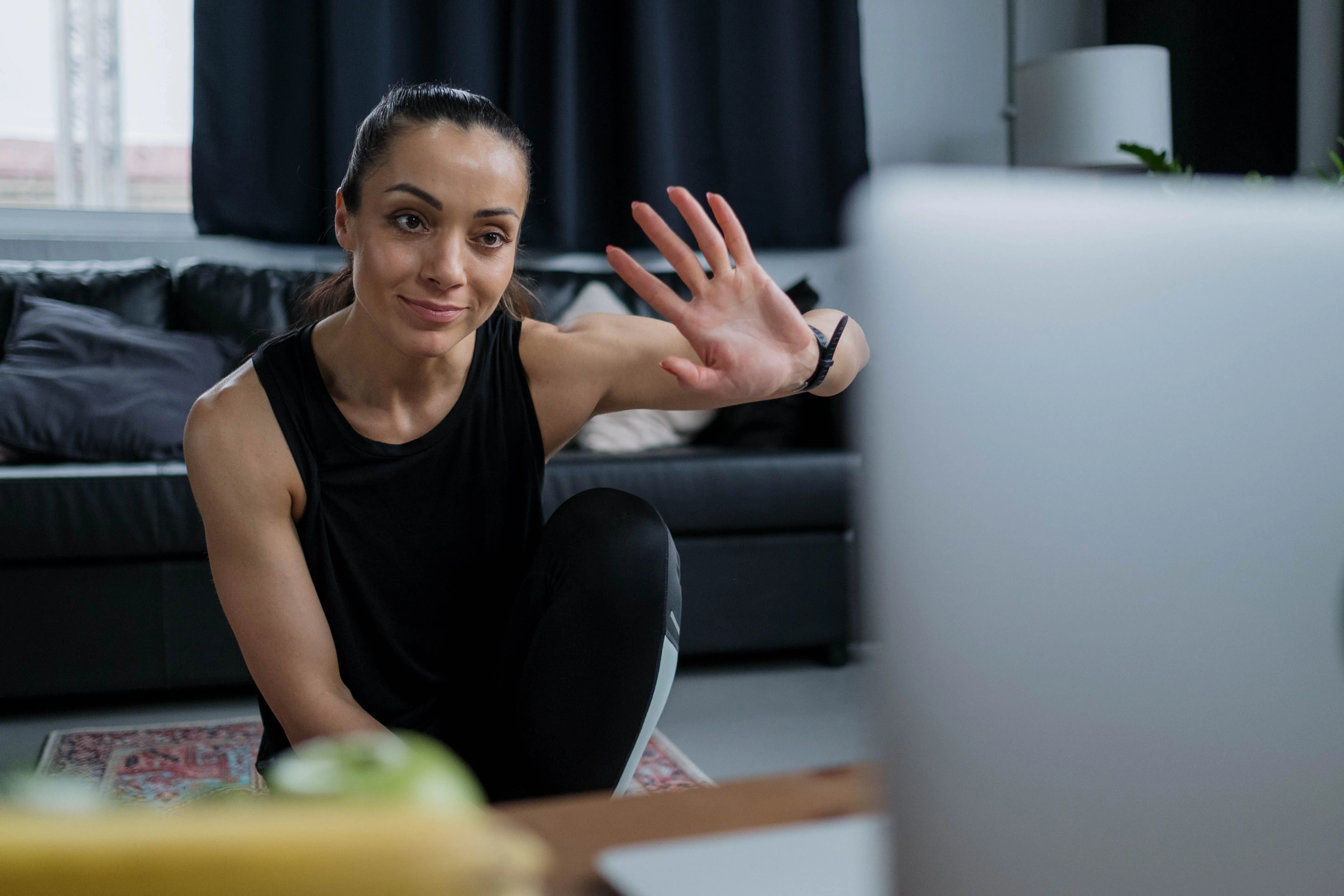 A woman in athletic clothing waves at a laptop during a video call in a modern living room, featuring a black sofa, potted plants, and soft natural lighting, creating a casual and inviting atmosphere.