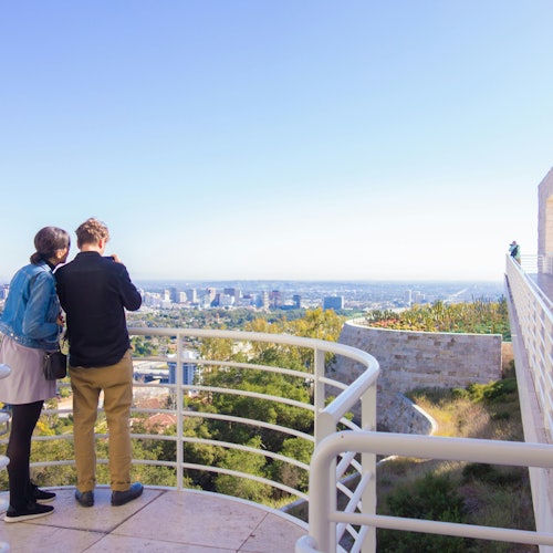 Two people stand on a terrace overlooking a cityscape, with railing and a clear blue sky in the background.