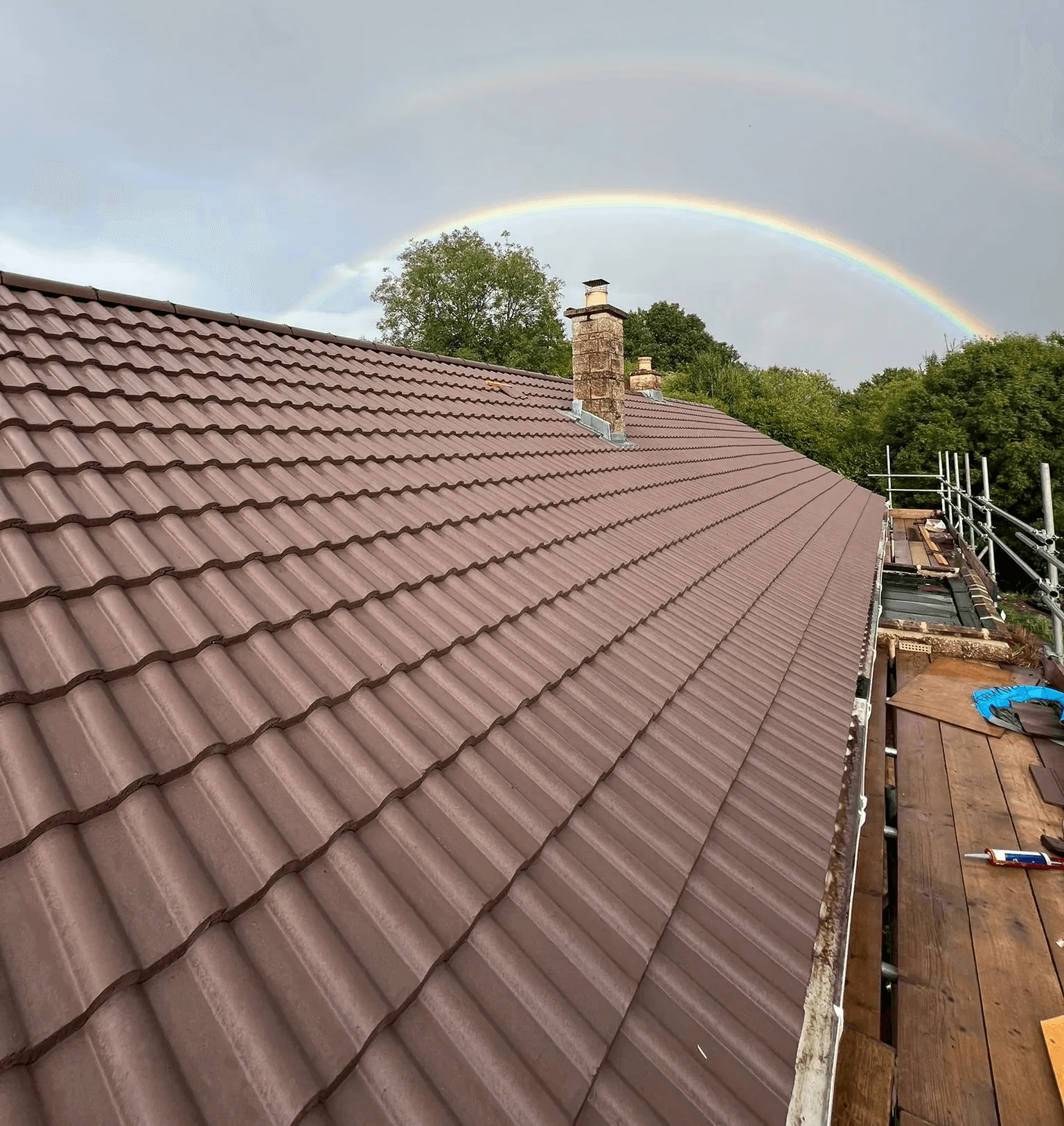 Brown tile roof with a chimney and a bright rainbow arching across the sky. The image is taken from the side.