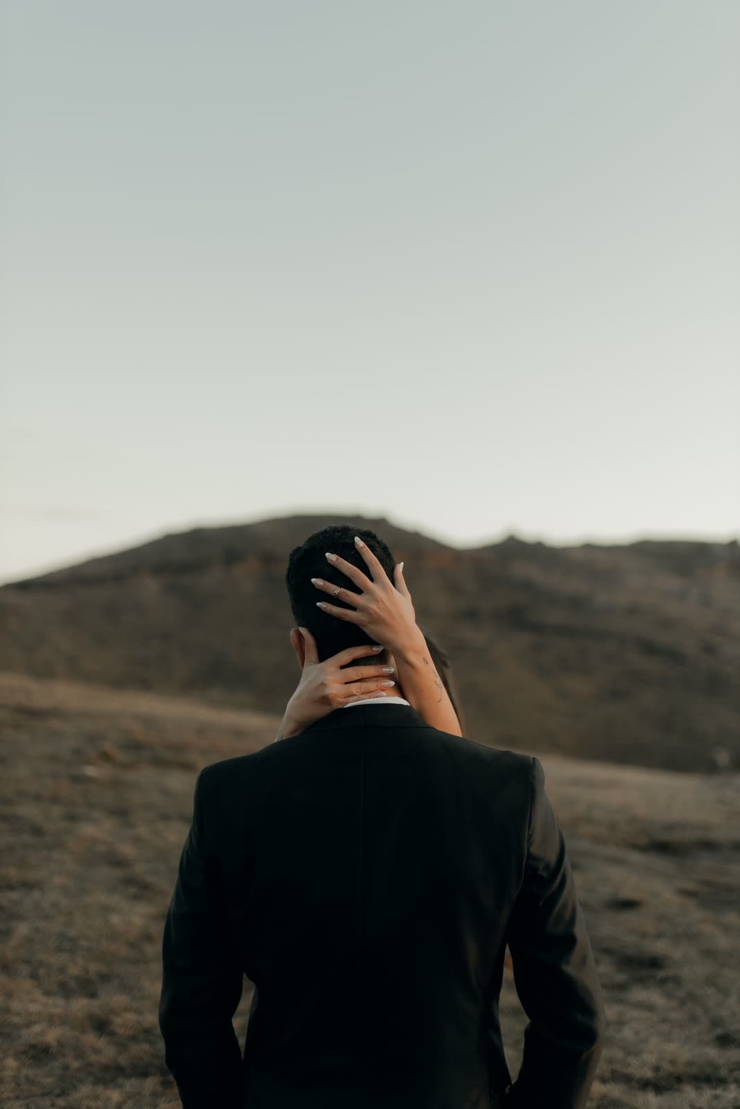 Groom seen from behind with the bride's hand on his neck against a dark hill