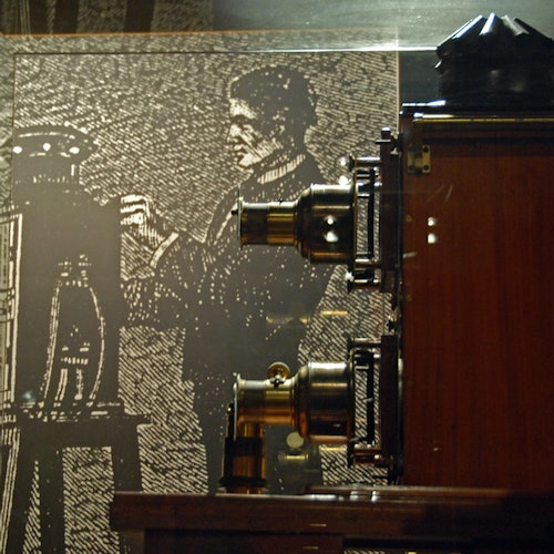 Vintage camera equipment displayed in a museum, with a background illustration of a man operating similar equipment.