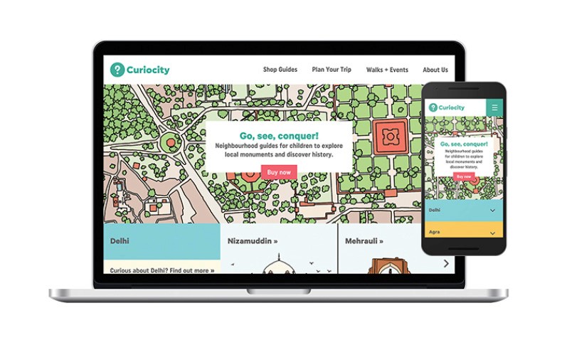 A laptop displays the Curiosity website, featuring a map of Delhi with a central red square and surrounding green areas. The website’s header includes “Curiosity” logo, navigation links (Shop Guides, Plan Your Trip, Walks + Events, About Us), and a “Go, see, conquer!” slogan. Below the map, a white box contains the text “Neighbourhood guides for children to explore local monuments and discover history.” A smaller smartphone screen shows the same website interface, emphasizing “Go, see, conquer!” and “Neighbourhood guides for children to explore local monuments and discover history.” The laptop and phone are positioned on a white surface against a white background.