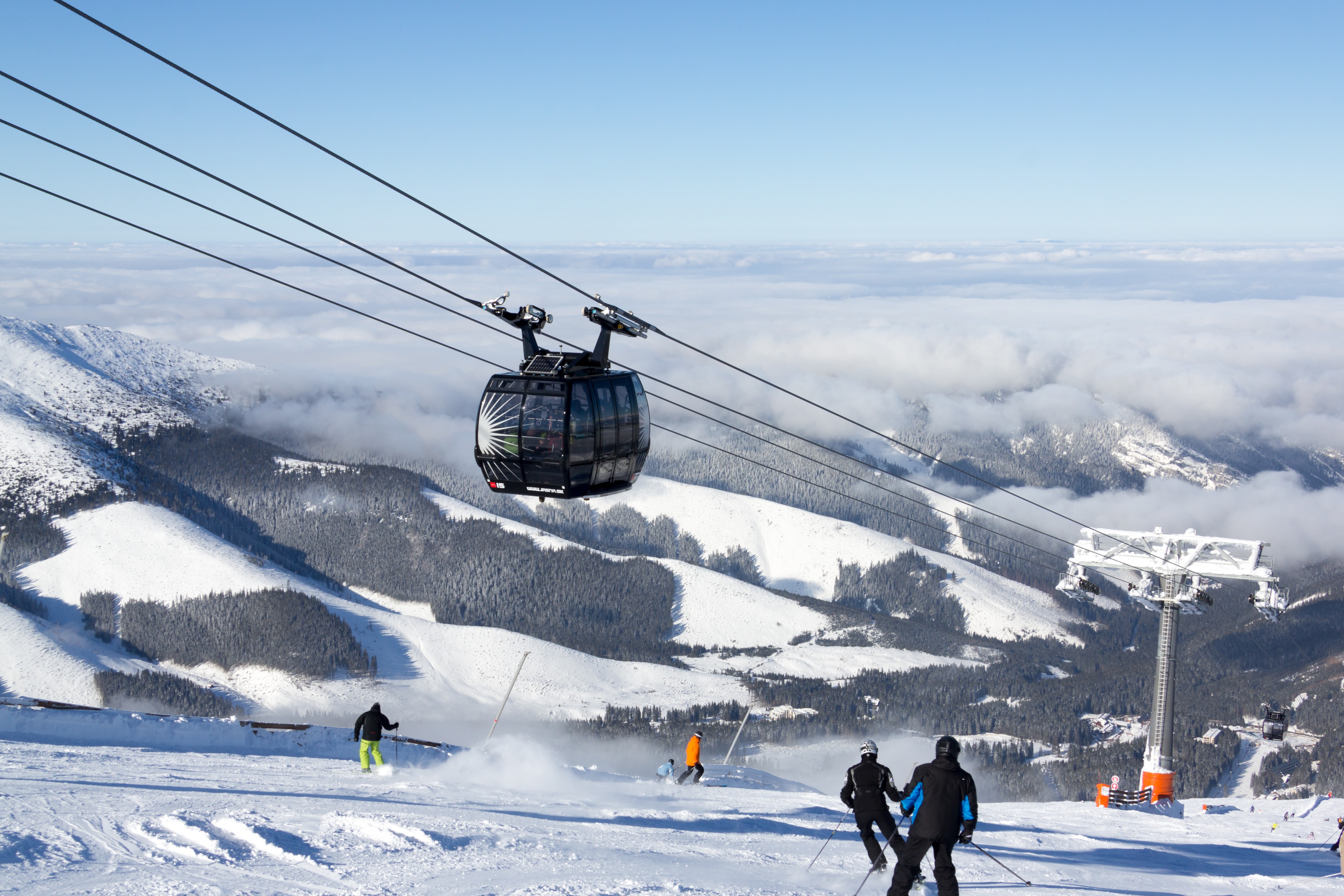 Skiers descend a snowy slope beneath a gondola gliding over a wide mountain landscape covered in winter clouds.