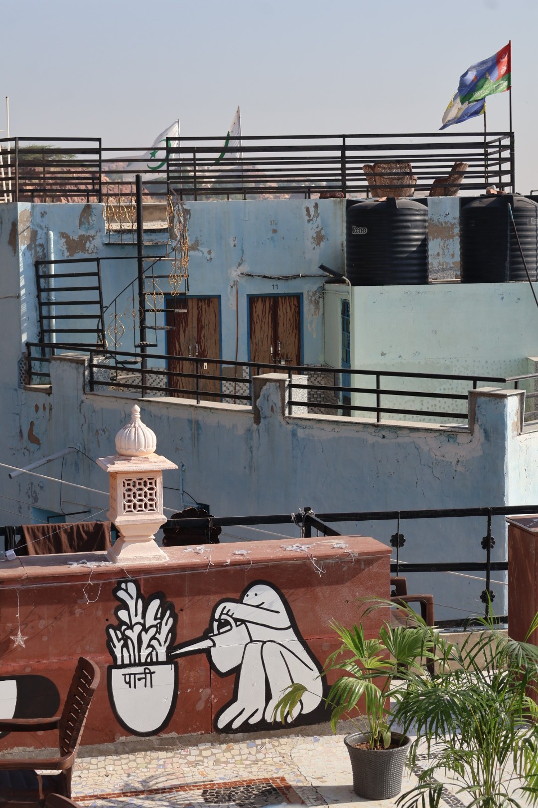 Mural painting of a joyful cartoon character watering a potted plant with the word ‘पानी’ written on it, painted black-and-white on a brown restaurant terrace with tables and oriental decorations visible.
