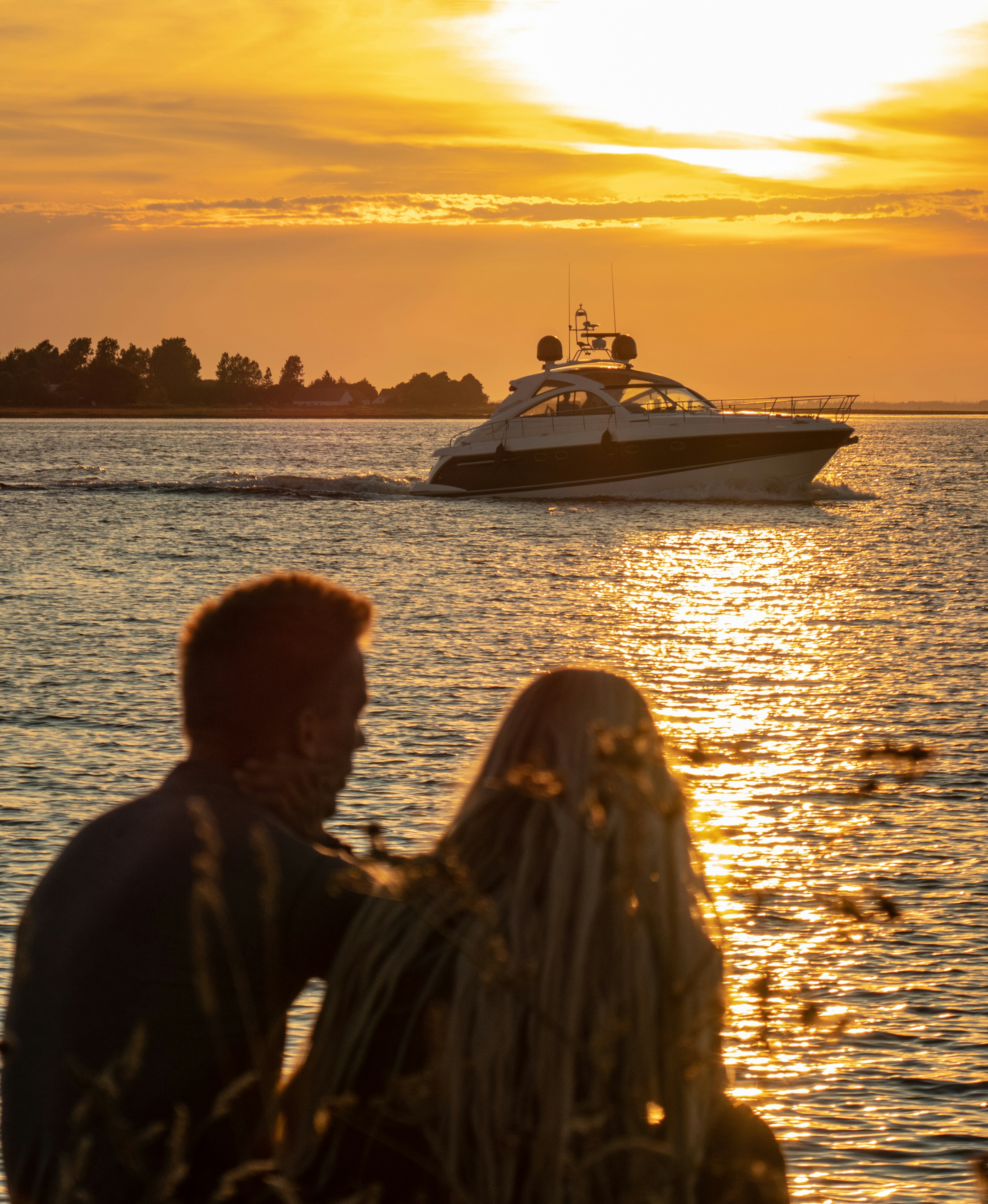 A couple sits by the shore, watching a luxurious yacht sail across the shimmering water at sunset, with golden hues reflecting off the calm sea.