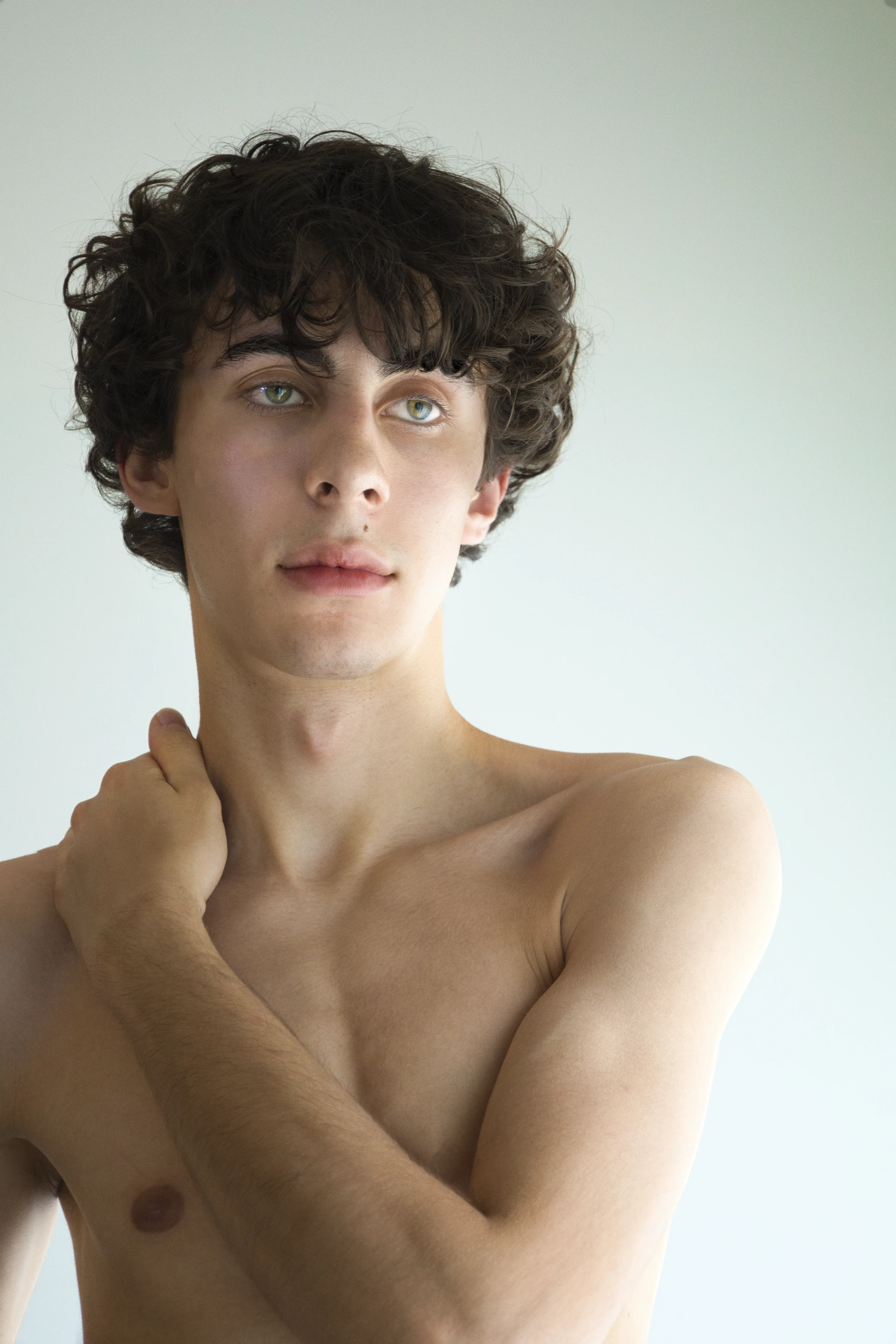 A shirtless young person stands against a light background, looking thoughtfully to the side.