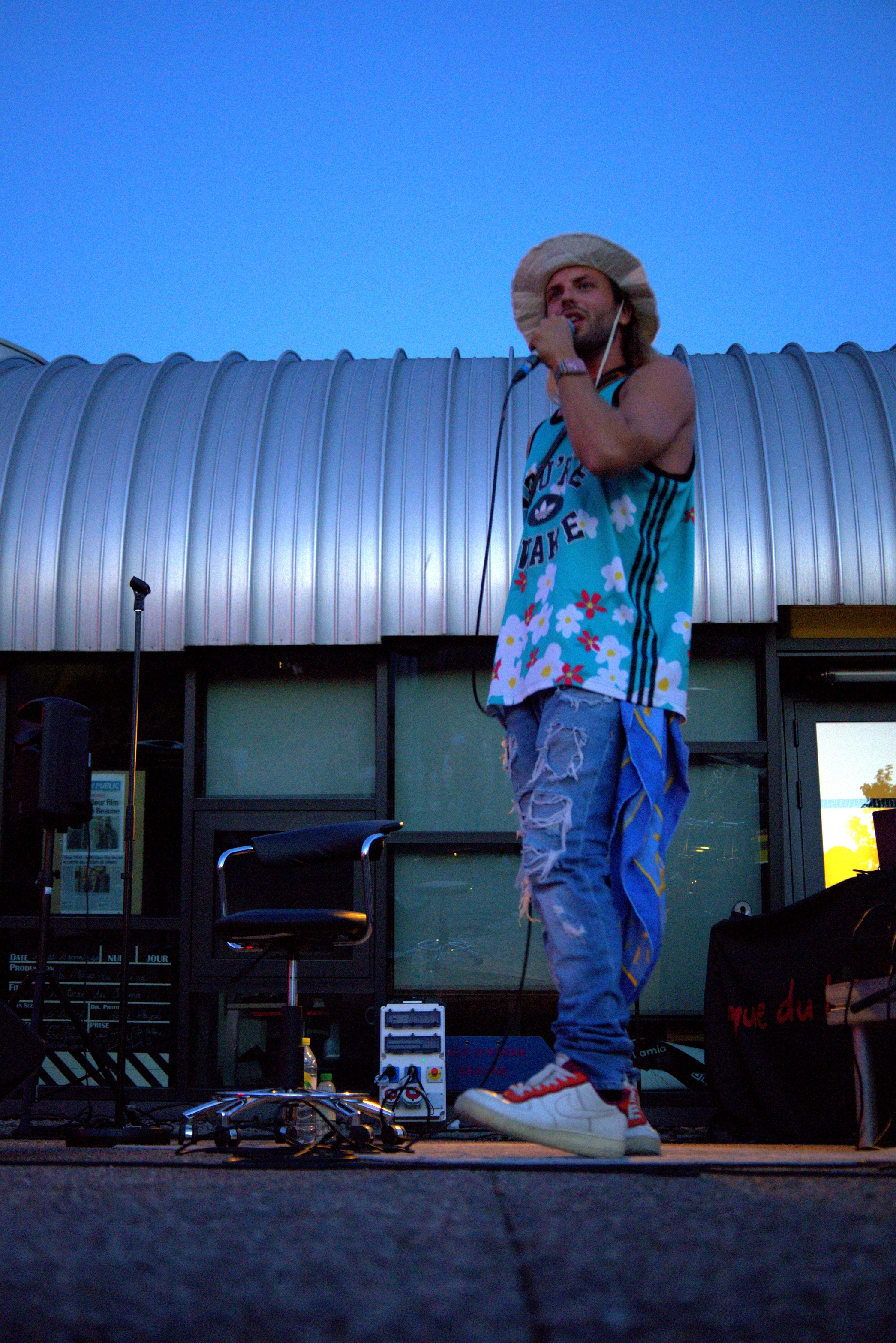 A person stands outside in casual attire, holding a phone, with a blue sky and building behind them.