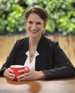 A person smiling while holding a cup, seated at a wooden table with greenery in the background.