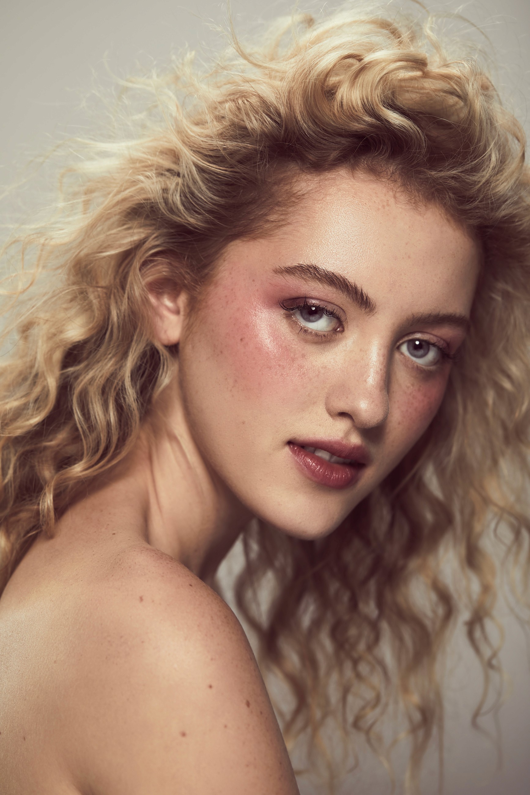 Beauty portrait of woman with voluminous blonde curly hair and freckled skin, featuring natural makeup with rose-toned blush and soft lighting against neutral background.