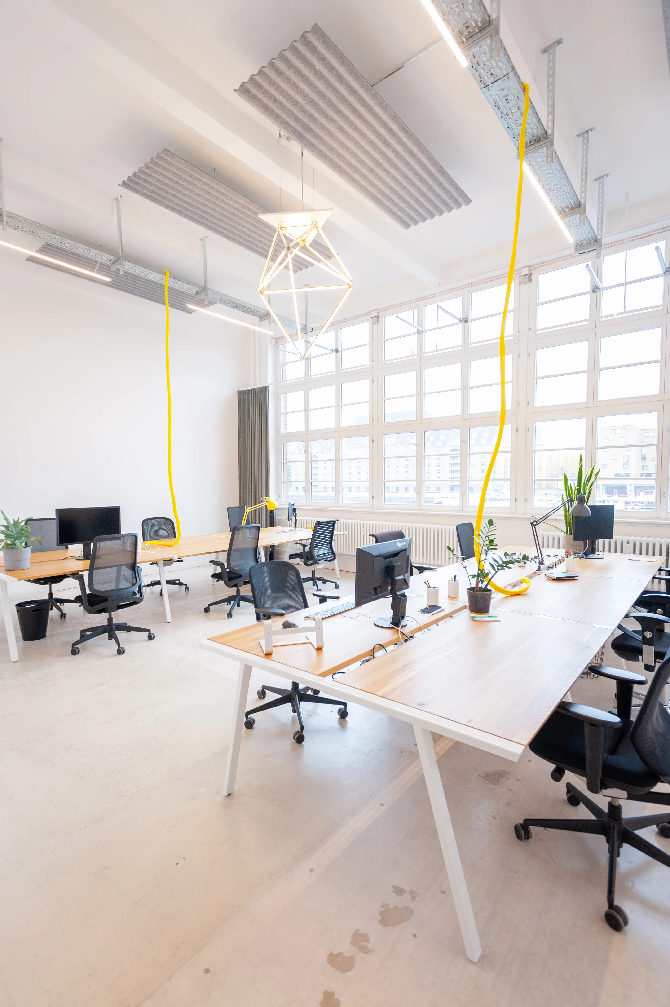 Open-space office at LA RED with white desks, black mesh chairs, large industrial windows, corrugated metal acoustic ceiling panels, yellow textile cables and a geometric wire pendant light.